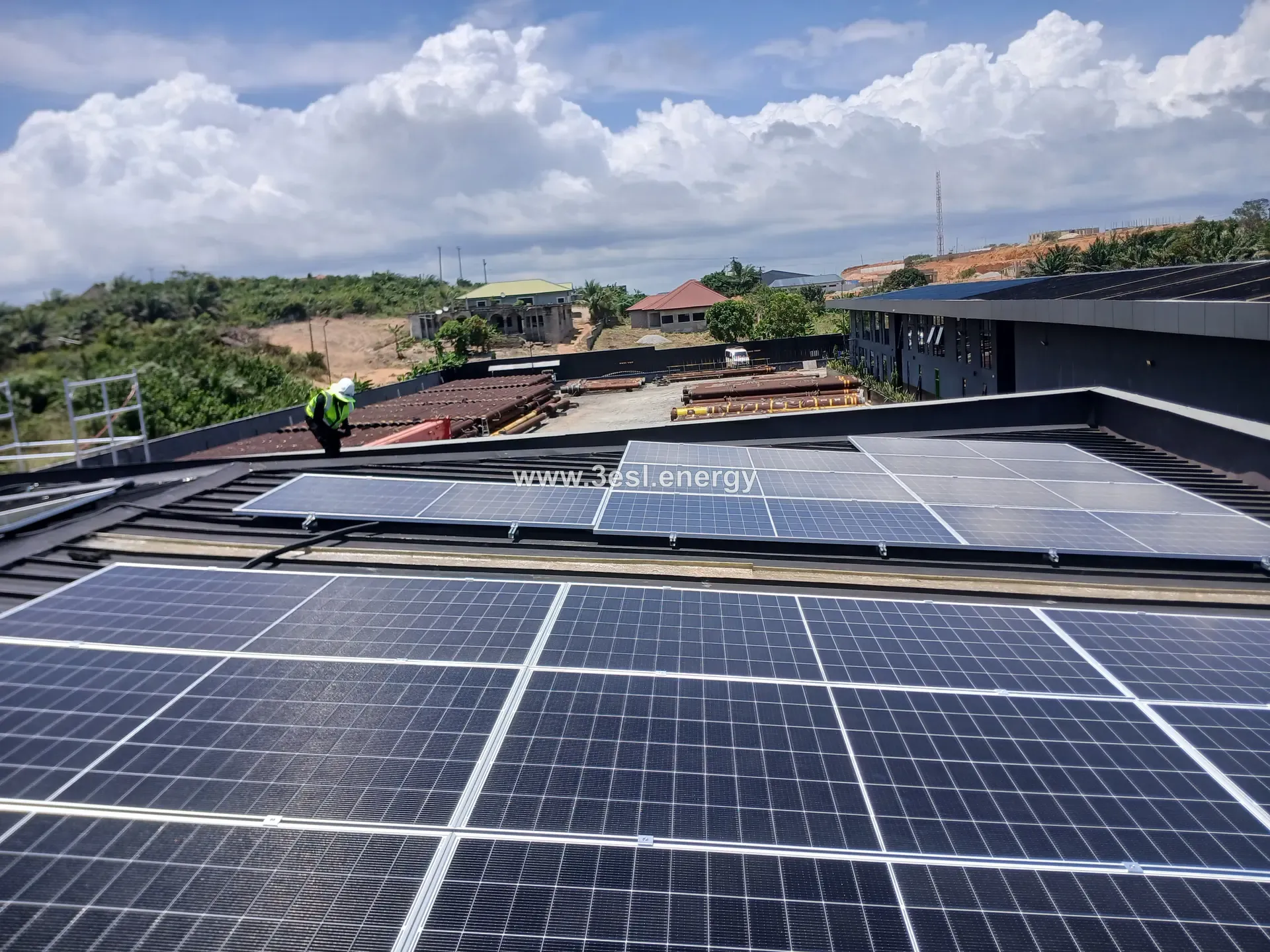 Rows of installed solar modules across a dark standing-seam commercial roof under a bright sky.